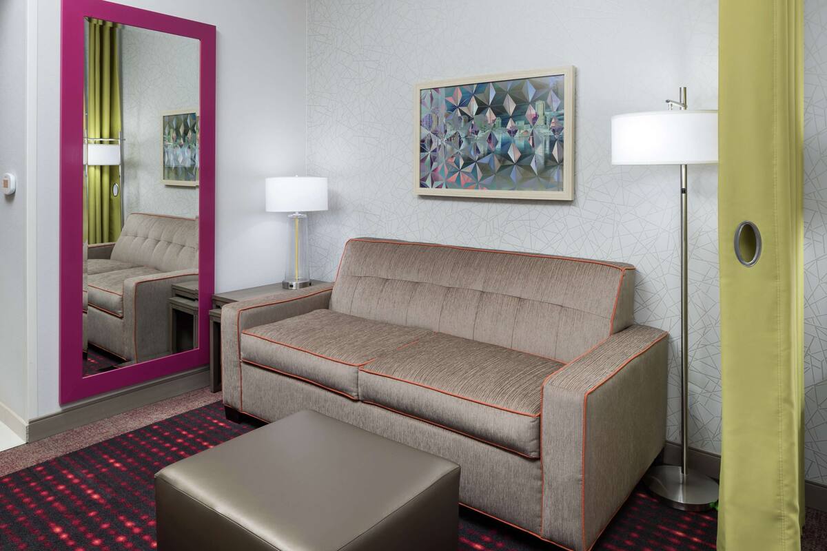 A hotel room with a sofa, a mirror, a painting, a lamp, and a carpet.