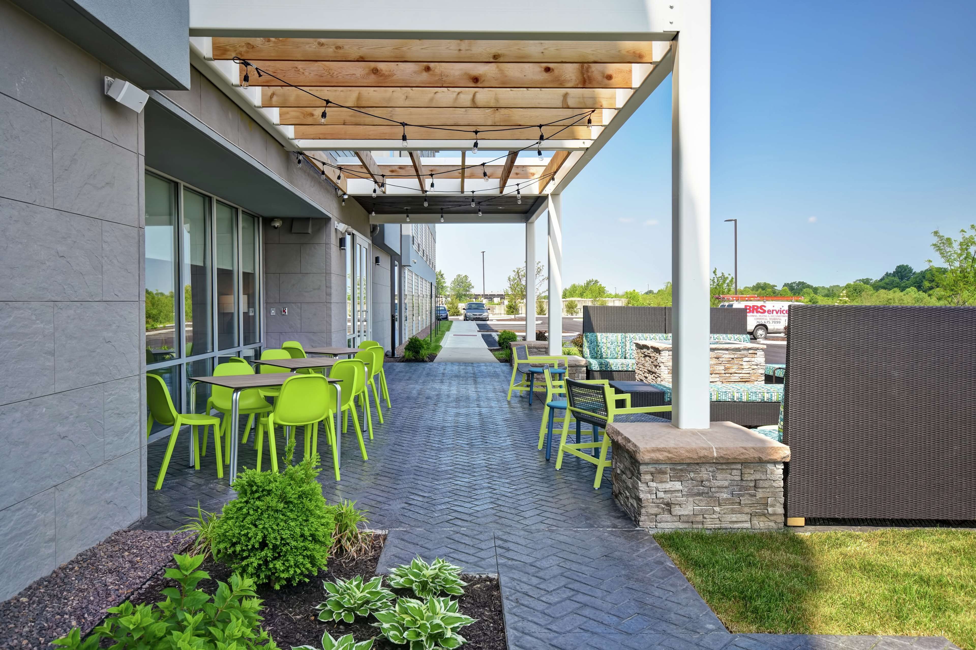 An outdoor patio area with green chairs and tables, a pergola with string lights, and a view of the street in the background.