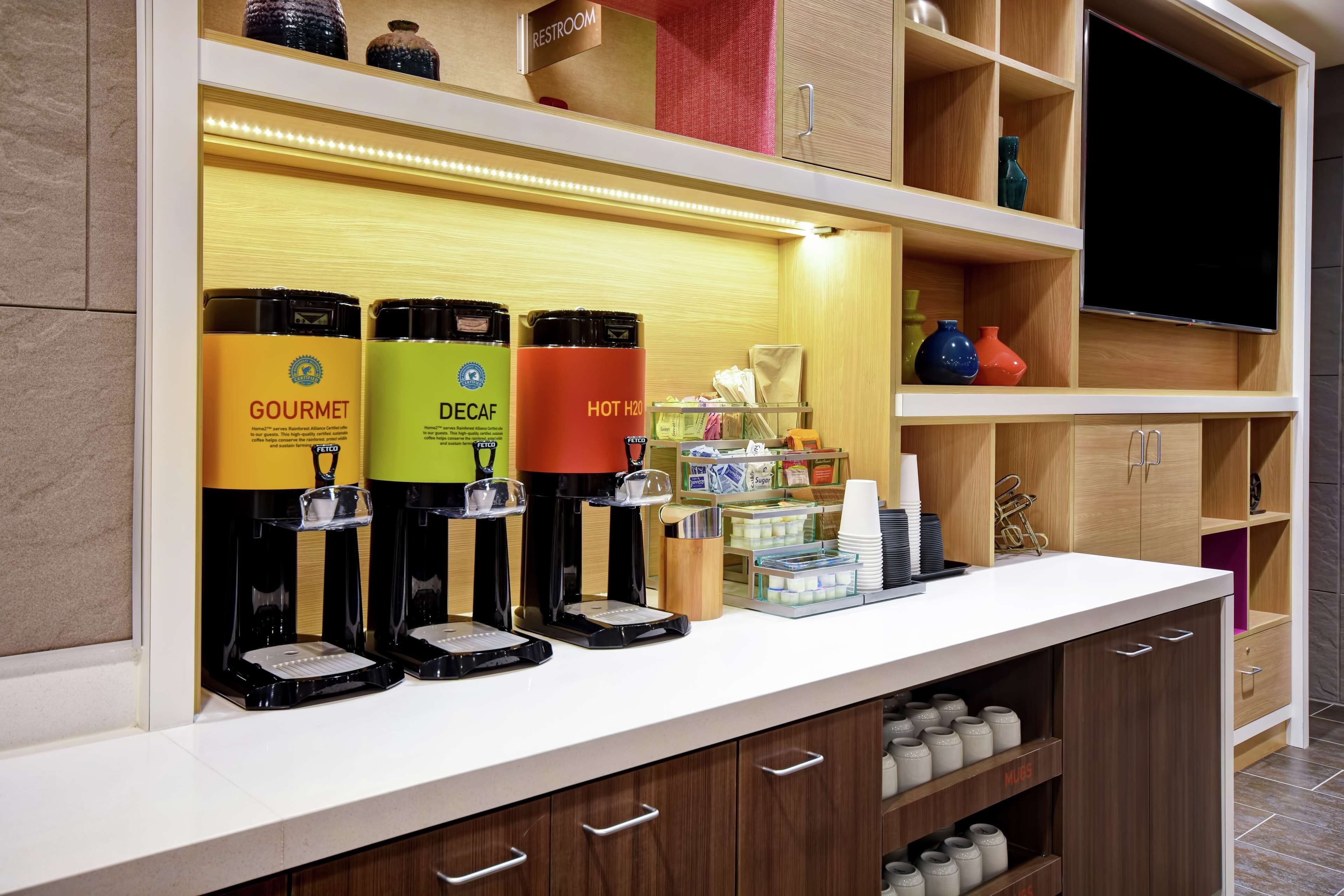 Una estación de café con tres máquinas de café identificadas como 'GOURMET', 'DECAF' y 'AGUA CALIENTE'. También hay estantes con diversos artículos, incluyendo un letrero de 'BAÑO', jarrones decorativos y un televisor.