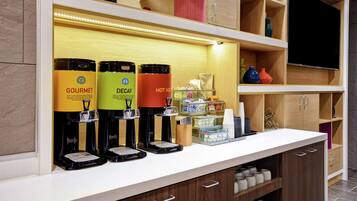 Una estación de café con tres máquinas de café identificadas como 'GOURMET', 'DECAF' y 'AGUA CALIENTE'. También hay estantes con diversos artículos, incluyendo un letrero de 'BAÑO', jarrones decorativos y un televisor.