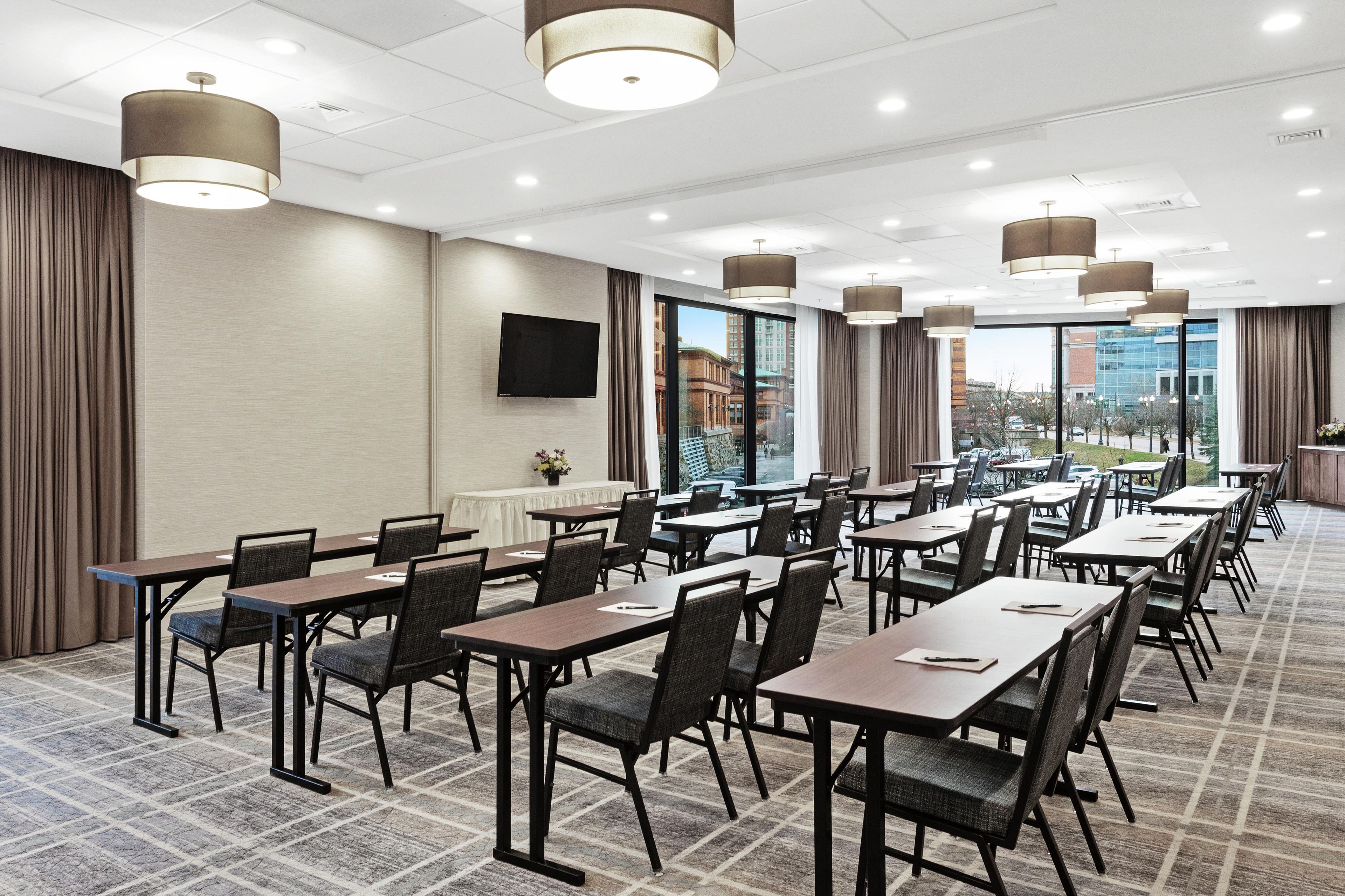A conference room with multiple tables and chairs, large windows with a city view, and modern pendant lighting.