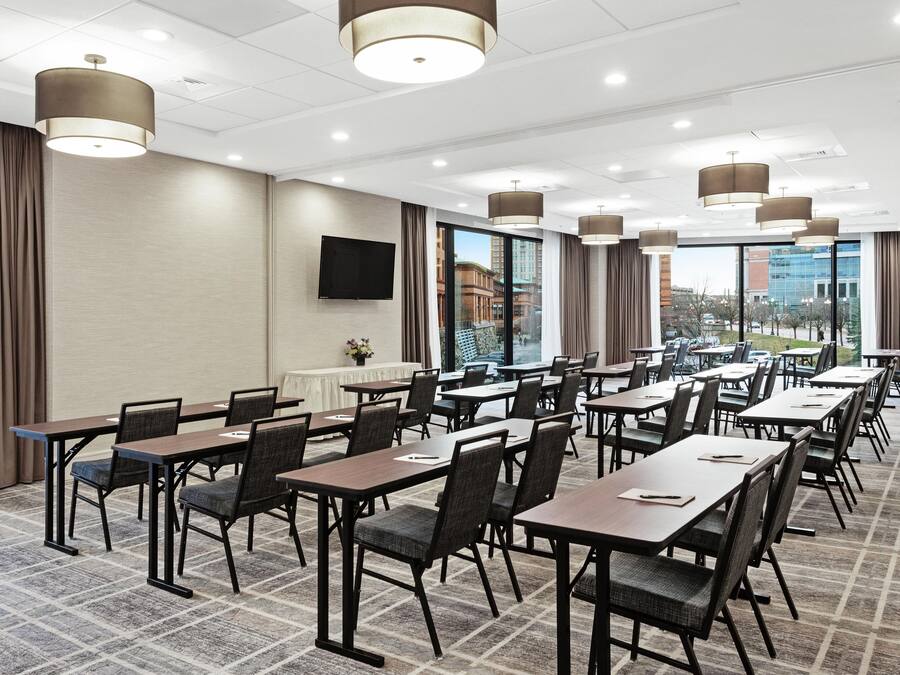 A conference room with multiple tables and chairs, large windows with a city view, and modern pendant lighting.