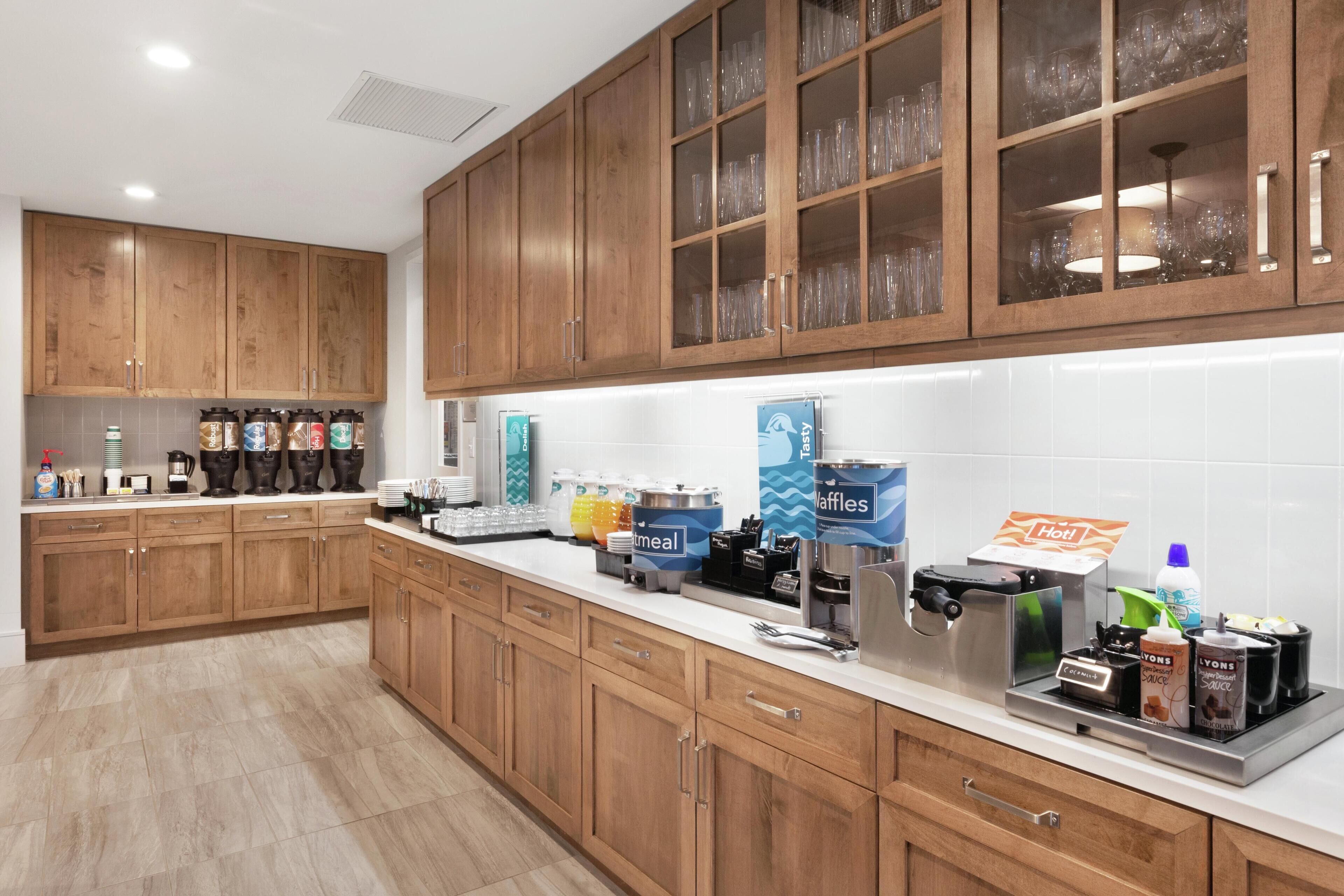 A modern kitchen with wooden cabinets, a stainless steel countertop, and various appliances including a coffee machine and waffle maker.