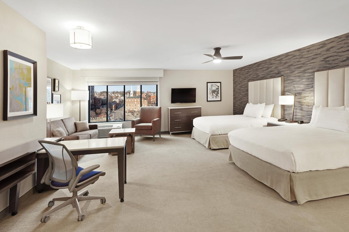 Une chambre d’hôtel équipée d’un bureau, de deux lits, d’une chaise, d’une télévision et d’une grande fenêtre donnant sur la ville.