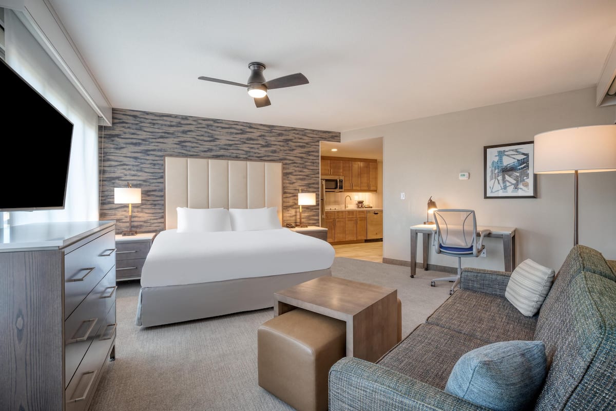 Une chambre d’hôtel avec un grand lit, un bureau, une chaise, un canapé et, au fond, une petite cuisine.