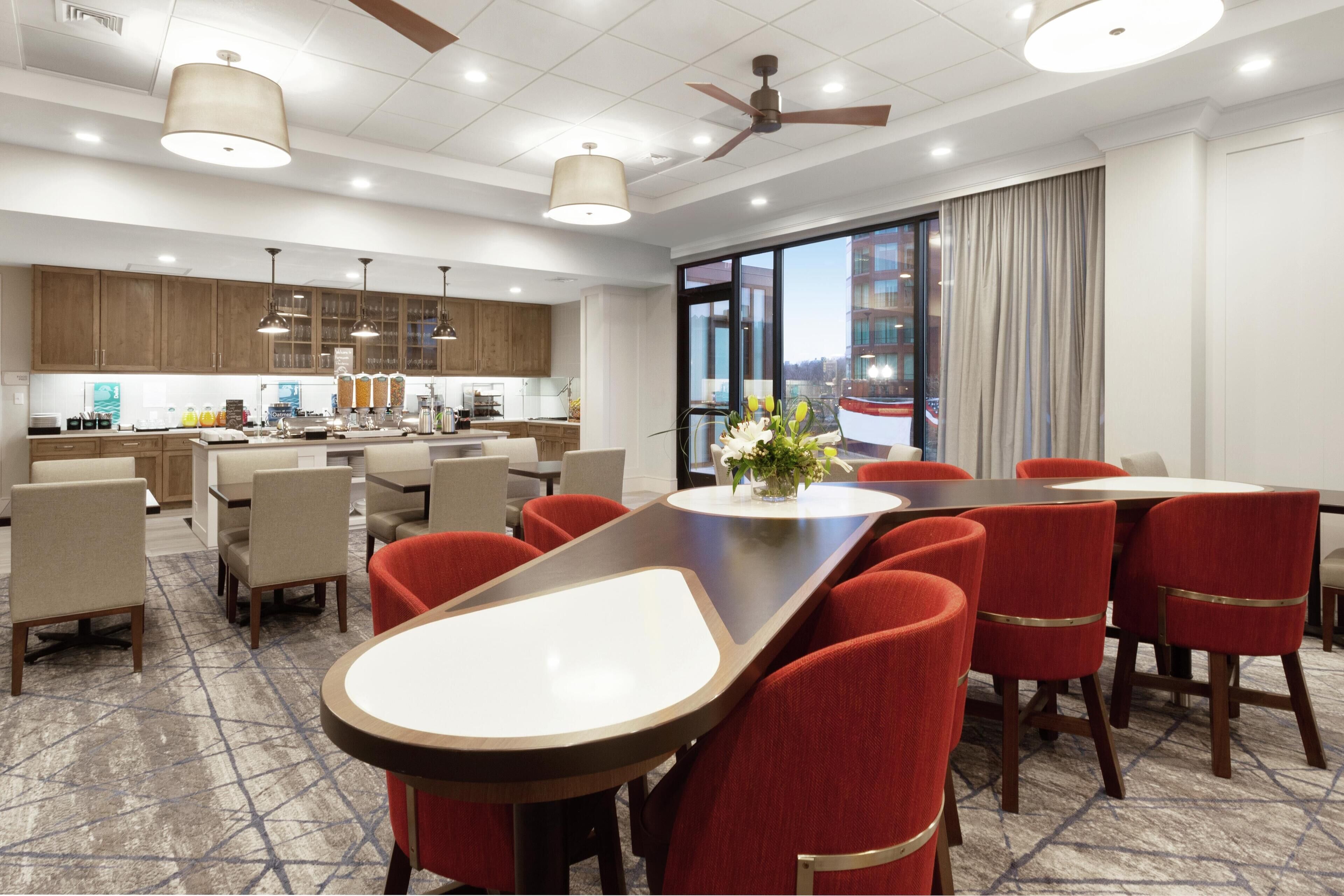 Ein moderner Hotel-Speisesaal mit einem großen Tisch, roten Stühlen und einer Küche im Hintergrund.