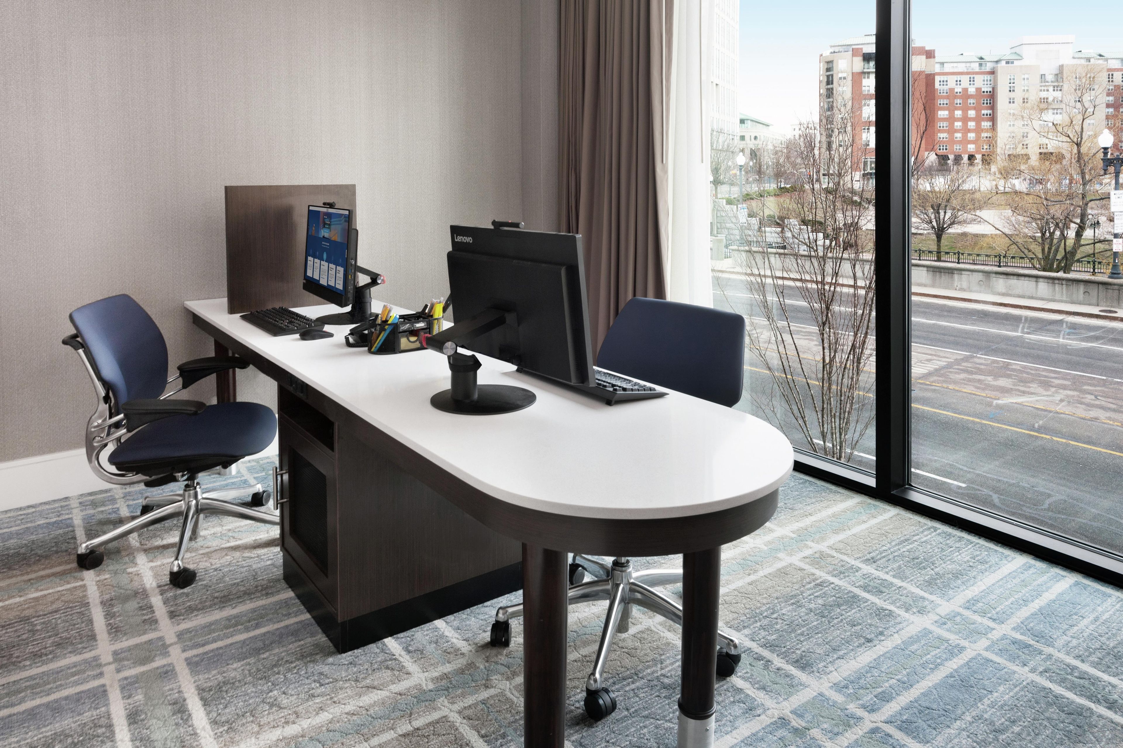 Habitación de hotel moderna con escritorio, dos sillas, una computadora y un ventanal con vistas a la ciudad.