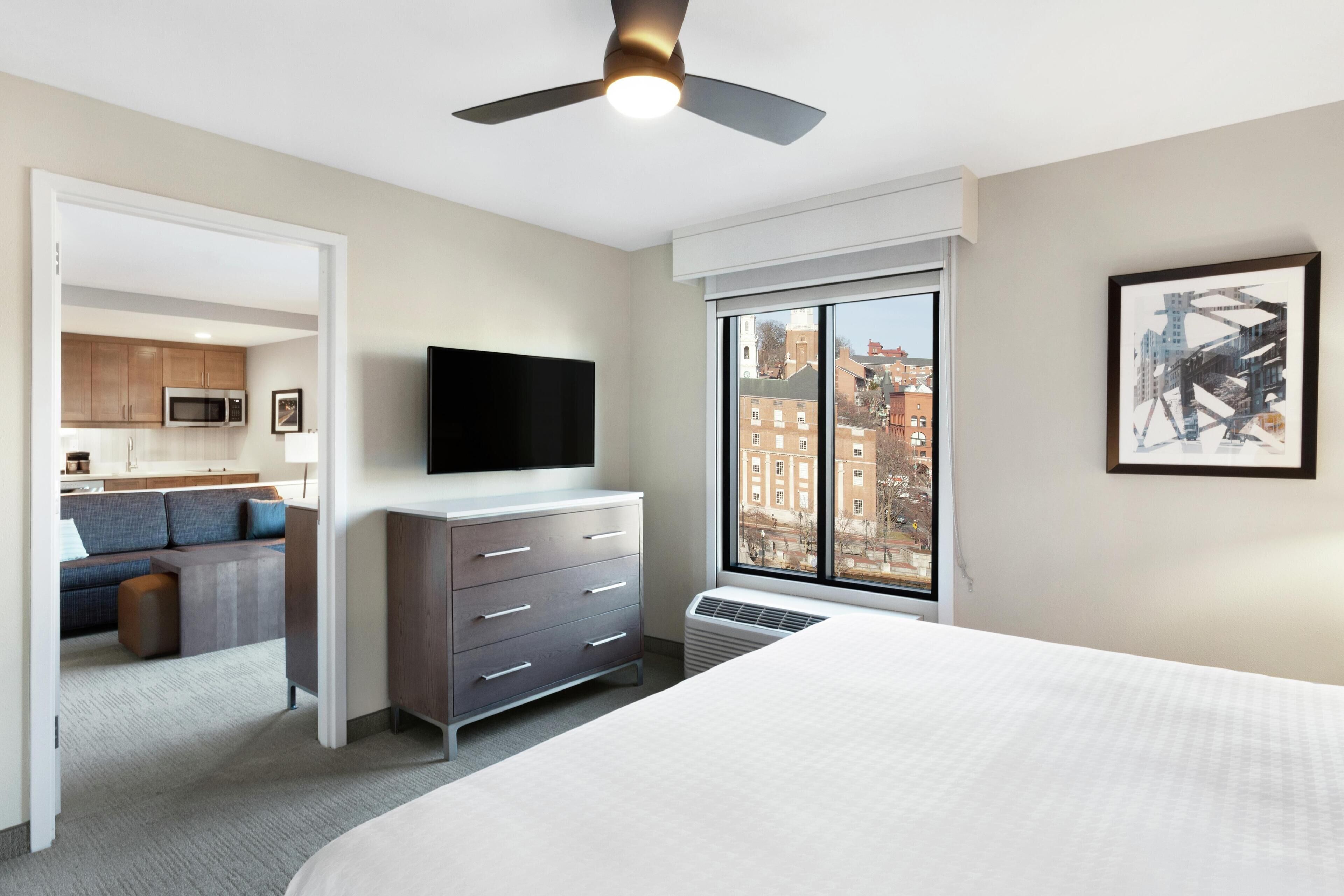 Habitación de hotel con cama, cómoda, televisor de pantalla plana, ventilador de techo, ventana grande con vista a la ciudad y una kitchenette al fondo.