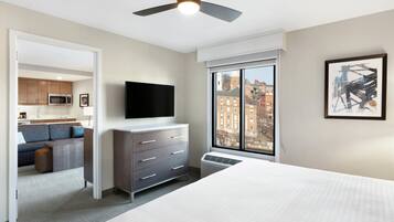 A hotel room with a bed, a dresser, a flat-screen TV, a ceiling fan, a large window with a city view, and a kitchenette in the background.