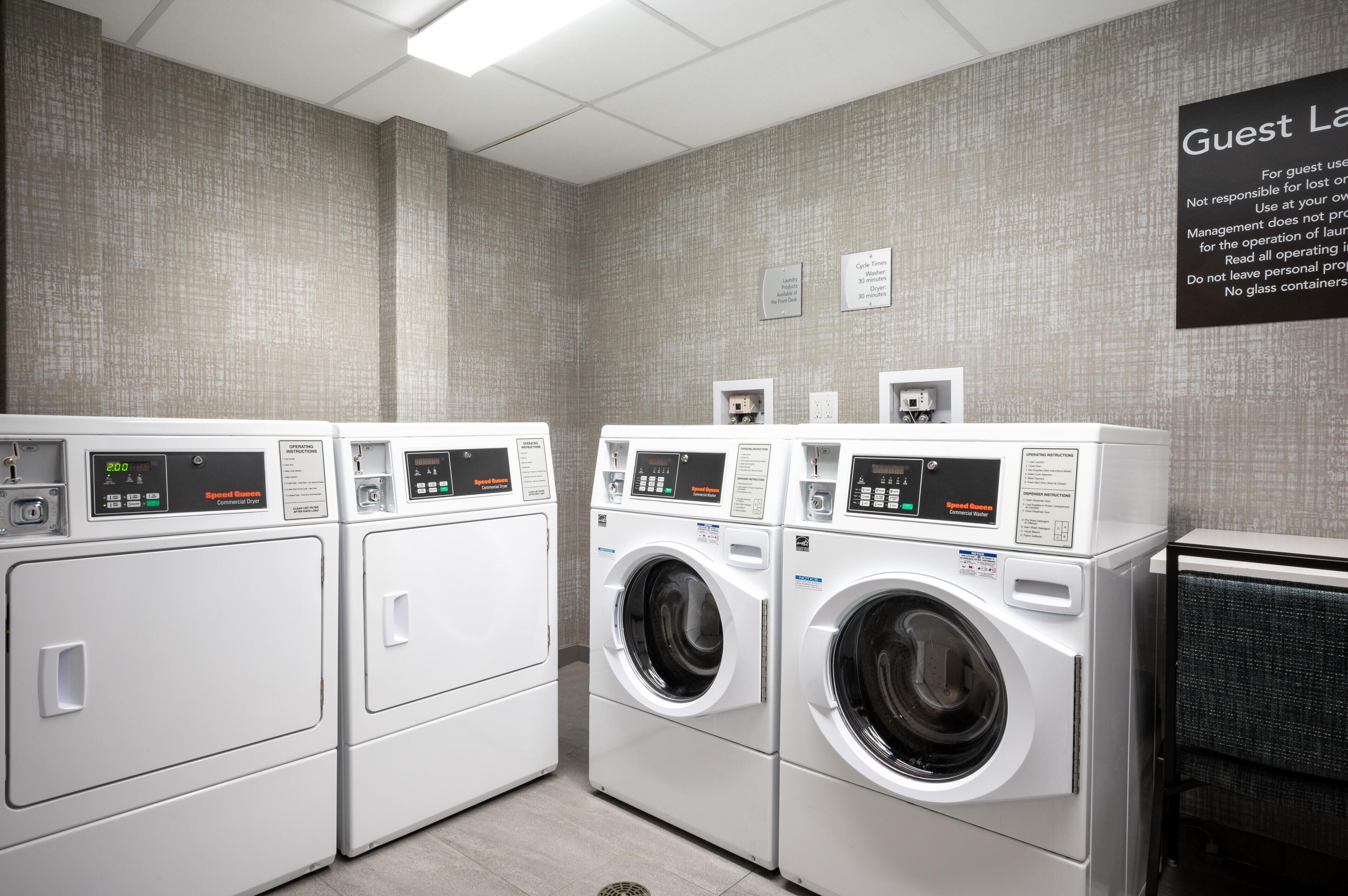 Een hotelwasserette met meerdere wasmachines en drogers, een bordje met gastenwasserijservices, en een wandbord met instructies.