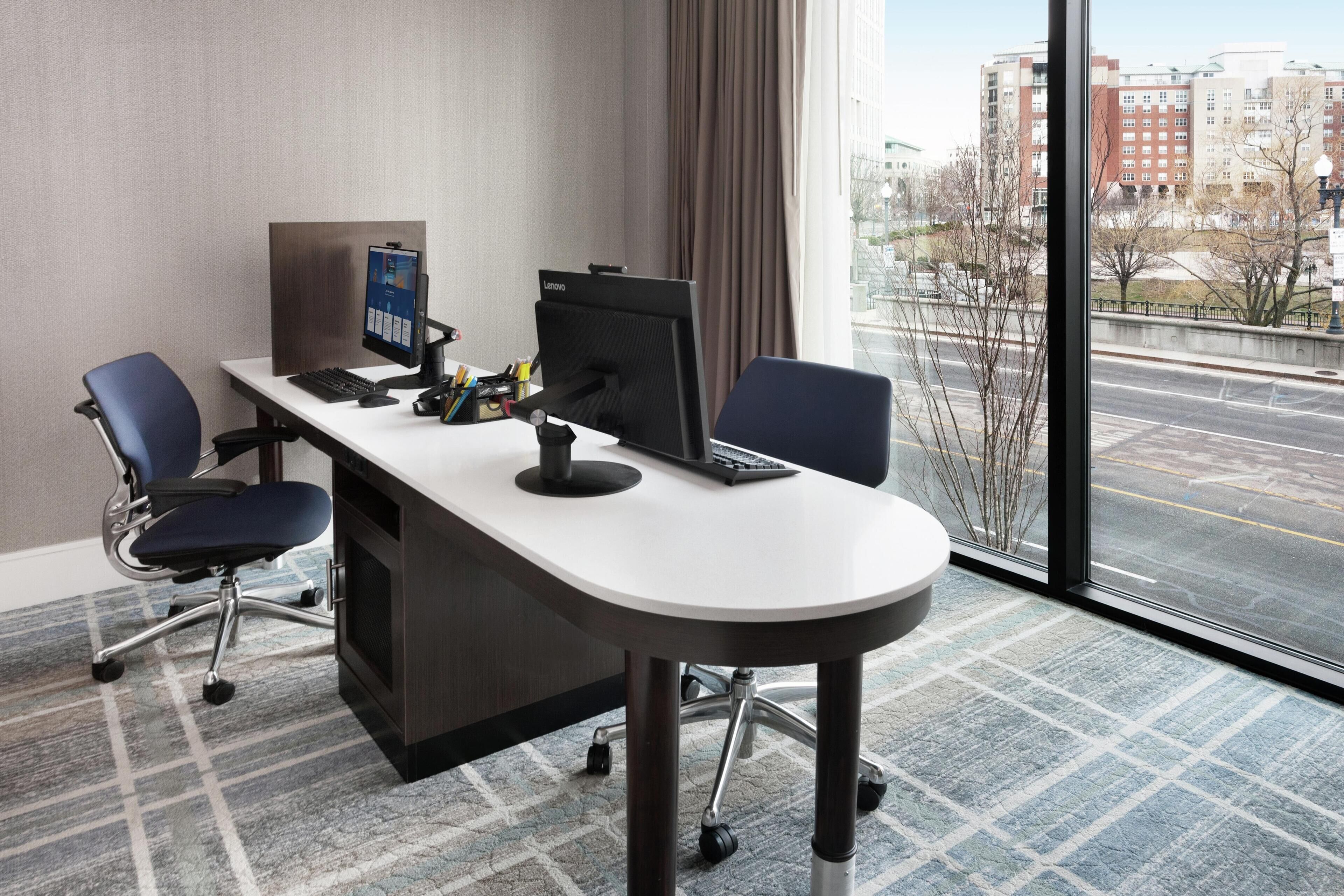 A modern hotel room with a desk, two chairs, a computer, and a large window offering a city view.