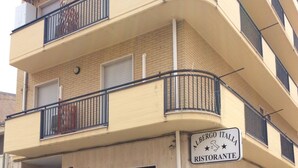 Un immeuble à plusieurs étages, avec une enseigne indiquant « Albergo Italia Ristorante » au rez-de-chaussée. Deux voitures sont garées devant, ainsi qu’un banc et un pot de fleurs près de l’entrée.