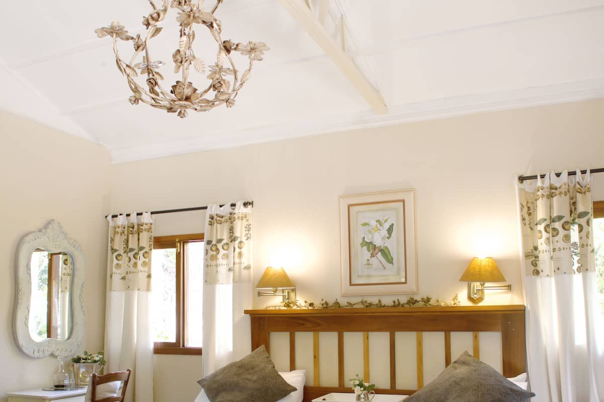 A bedroom with two beds, a wooden floor, a chandelier, a bedside table with a lamp, a mirror, and a window with curtains.