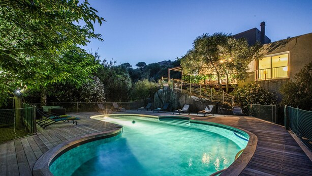 Une piscine avec une terrasse en bois, entourée d’arbres et bordée d’un bâtiment dont une fenêtre est illuminée.