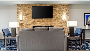 A modern hotel room with a large flat-screen TV mounted on a textured wall, a grey sofa, two side tables with lamps, and a patterned rug. - Comfort Suites Humble Houston IAH (Humble)