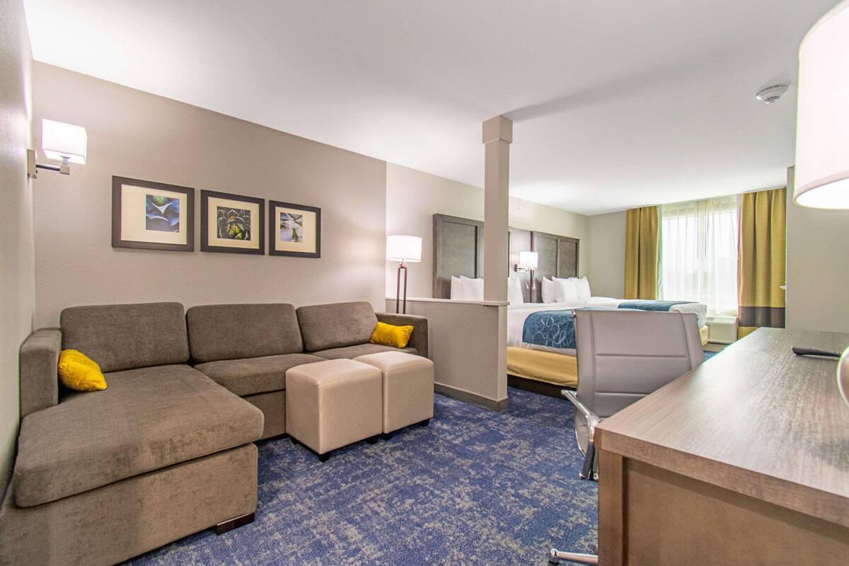 Een hotelkamer met een bank, een voetenbankje, een bureau en een bed met hoofdbord. Er hangen afbeeldingen in lijsten aan de muur en er is een raam met gordijnen.