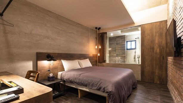Quarto moderno com uma cama grande, uma mesinha de cabeceira com abajur e vista para um banheiro através de uma porta de vidro.