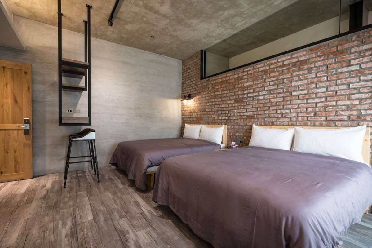 A bedroom with two beds, a wooden wardrobe, a metal ladder, and a brick wall.
