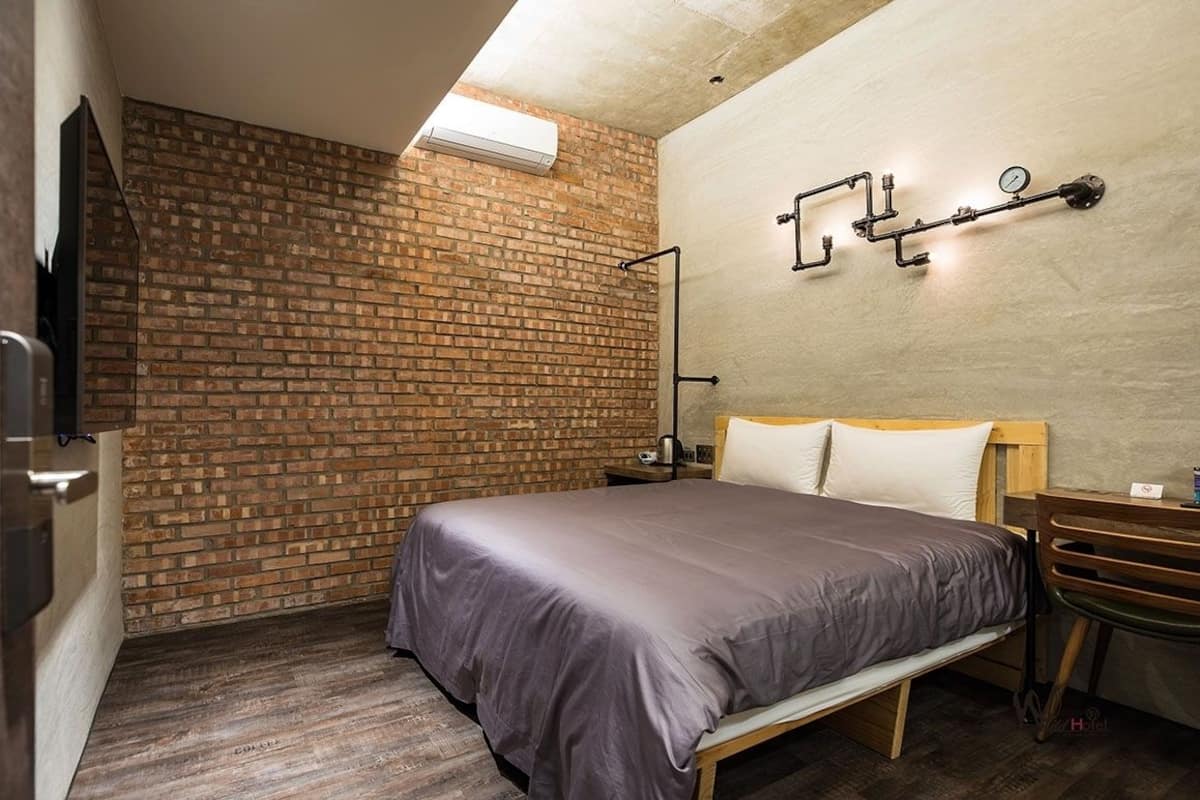 A bedroom with a brick wall, a bed with a wooden headboard, a nightstand, a desk with a chair, and a wall-mounted light fixture.
