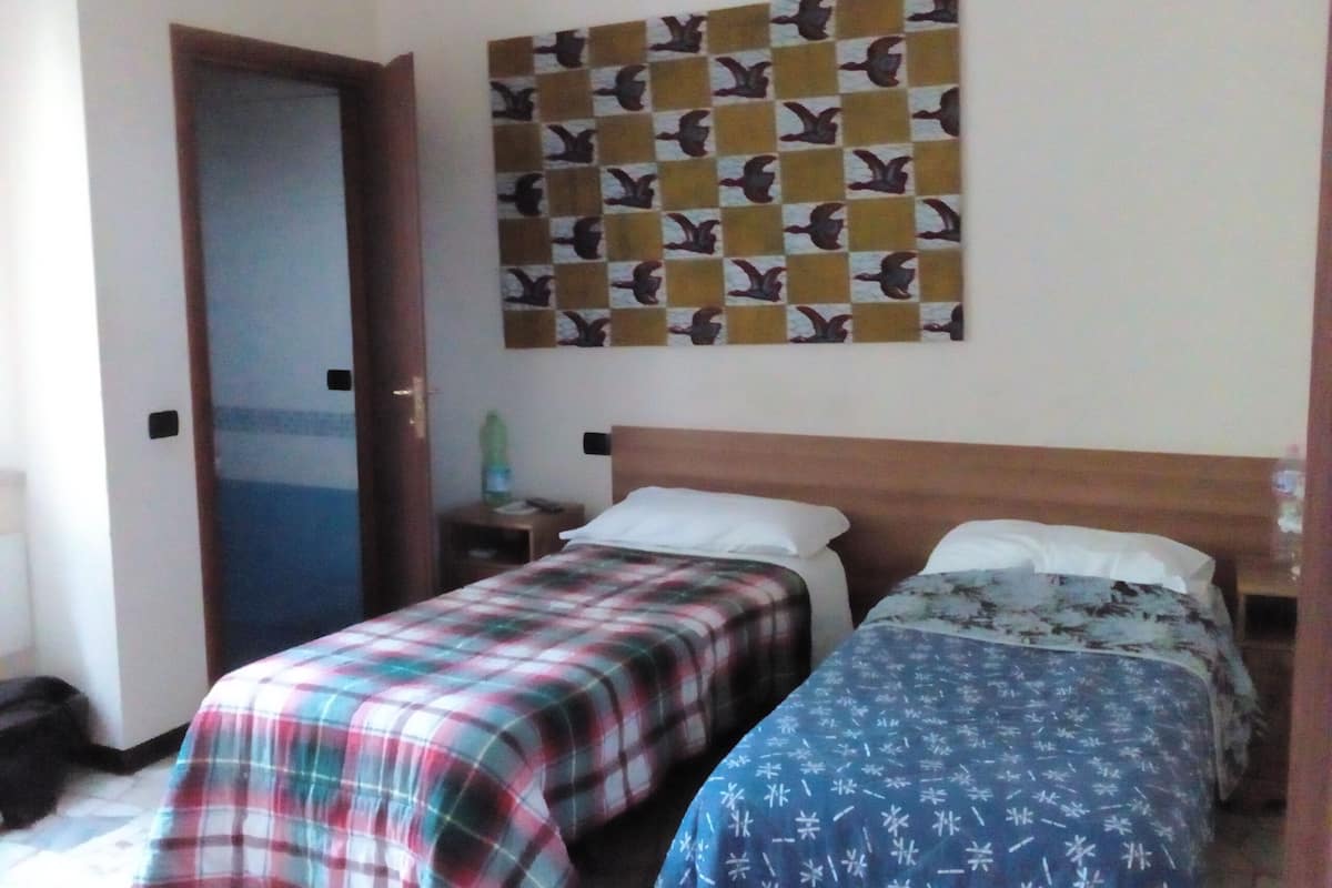 Ein Zimmer mit zwei Betten, einem Holzkopfstück, einer Wand mit Vogelmuster, einer Tür und einem kleinen Tisch mit einer Flasche.