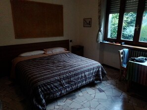 Et soveværelse med seng, natbord, stol, et vindue med udsigt til grøn vegetation og en radiator.