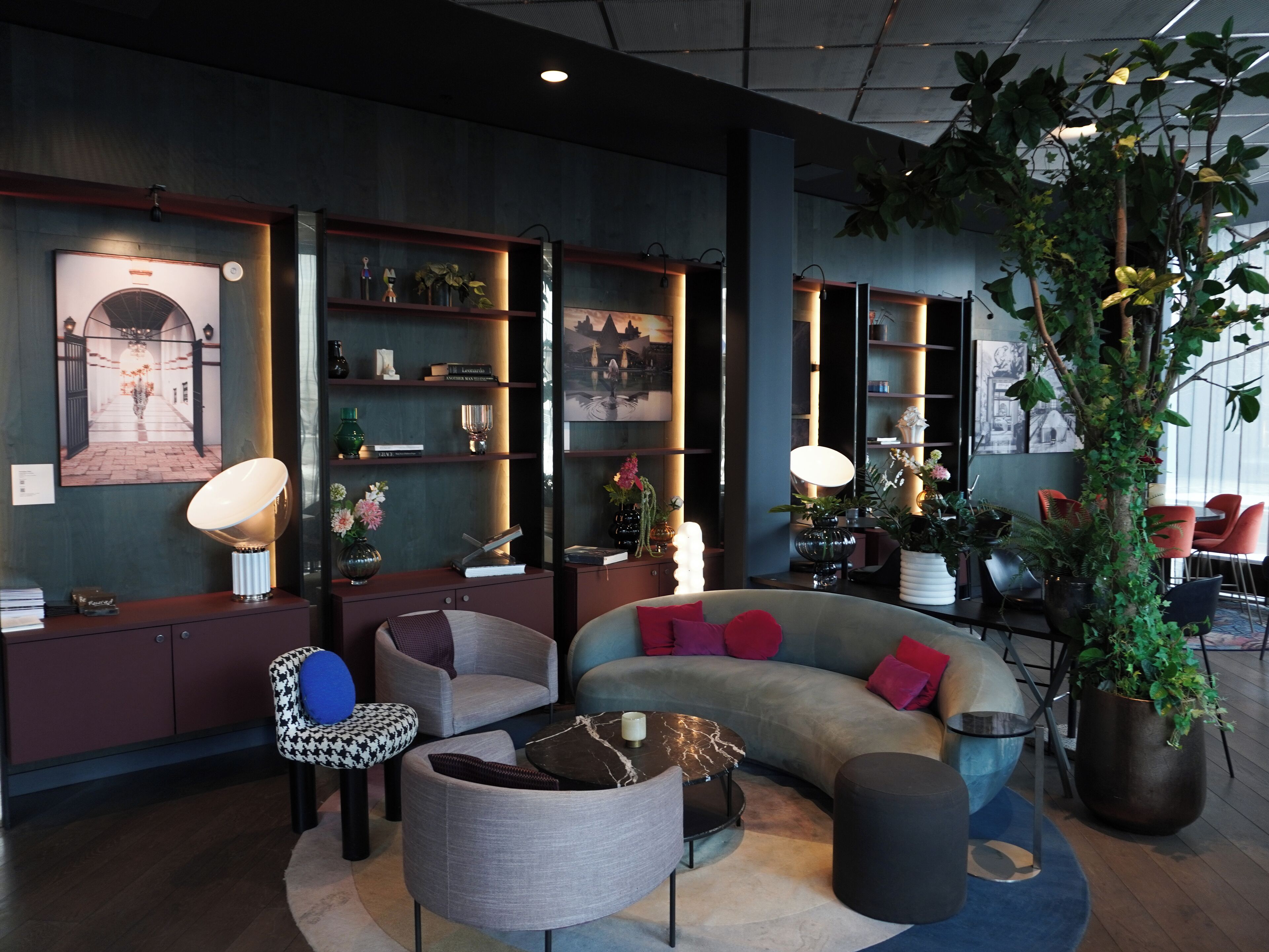 A modern interior with a curved sofa, armchairs, and a coffee table. There are shelves with decorative items and books. A large potted plant is in the corner.