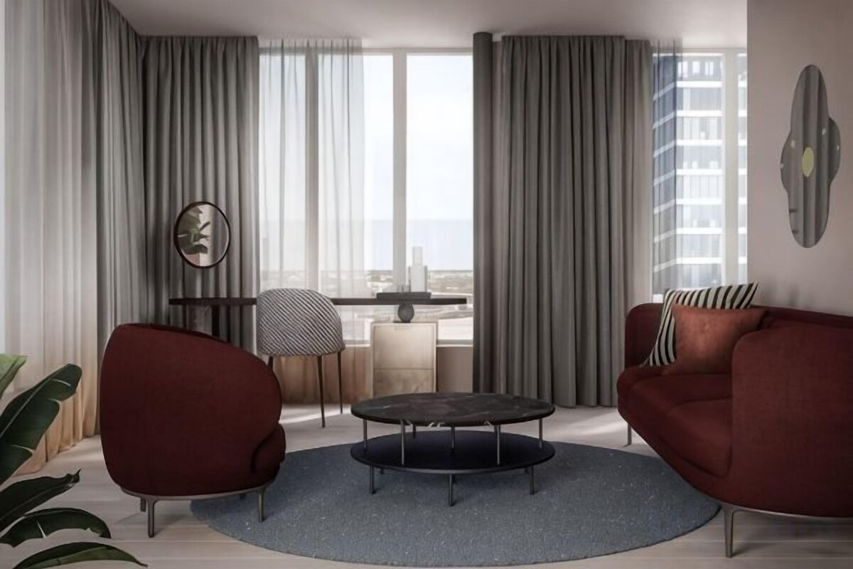 Una sala de estar moderna con una mesa redonda, dos sillones, un sofá y vistas a la ciudad a través de amplios ventanales.