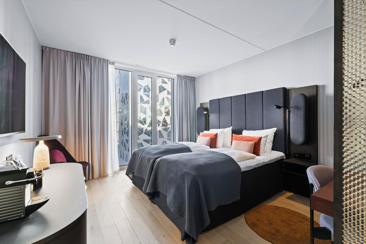 A modern hotel bedroom with a large bed, a desk, and a view of the city through large windows.