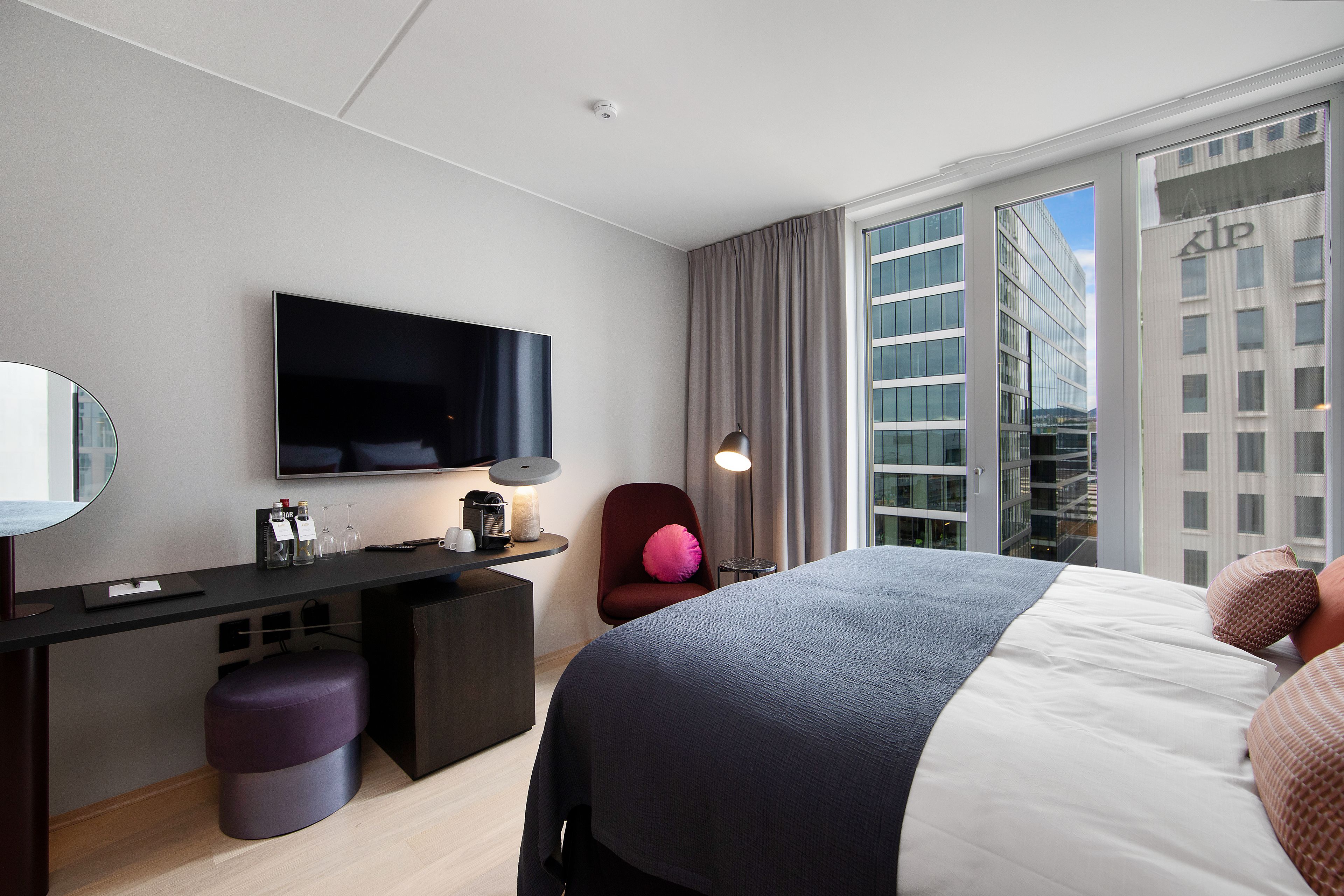 A modern hotel room with a large bed, a desk with a flat-screen TV, a chair, and a view of the city through large windows.
