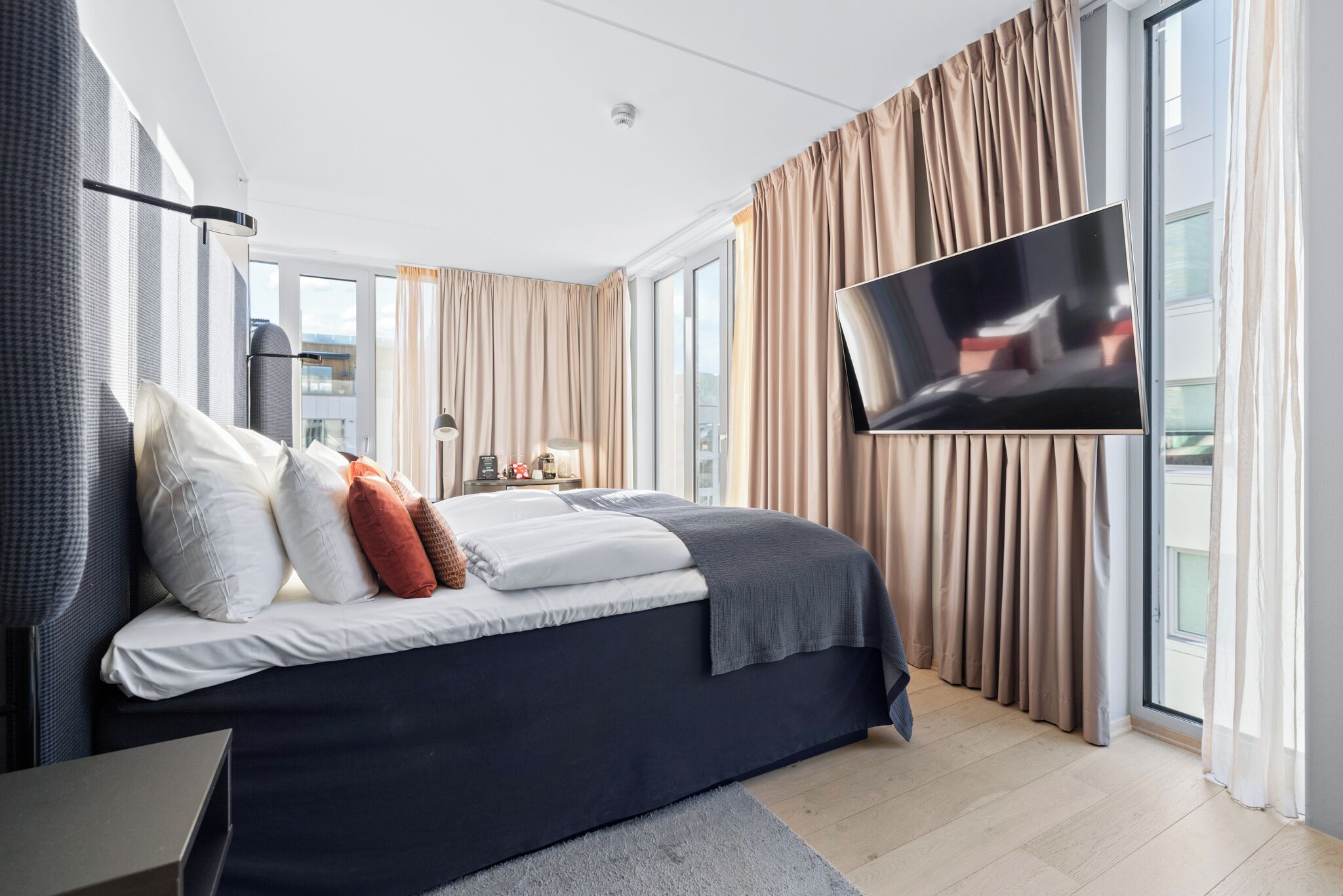 A modern hotel room with a large bed, a flat-screen TV, and a view of the city through large windows.