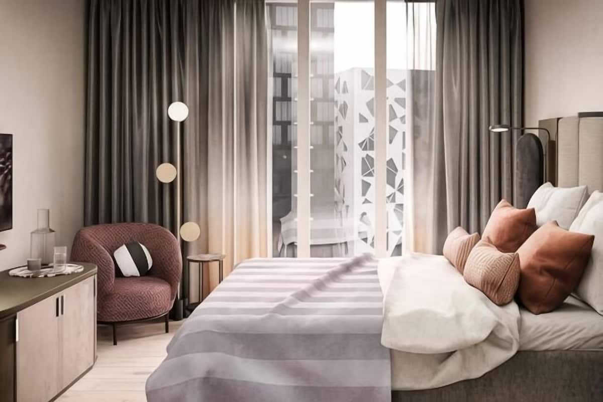 A modern hotel room with a large bed, a desk, a chair, and a view of the cityscape through the window.