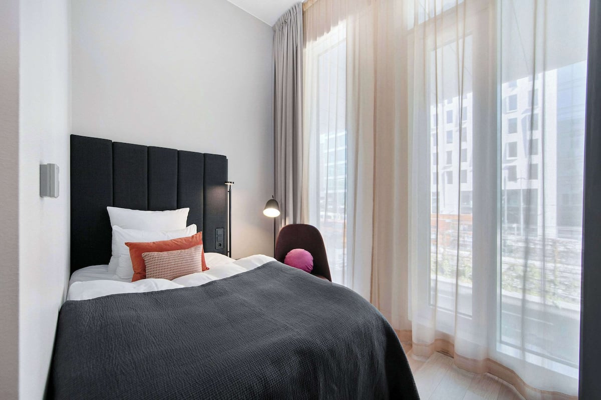 A modern hotel room with a large bed, a headboard, a bedside table with a lamp, and a view of the city through large windows.
