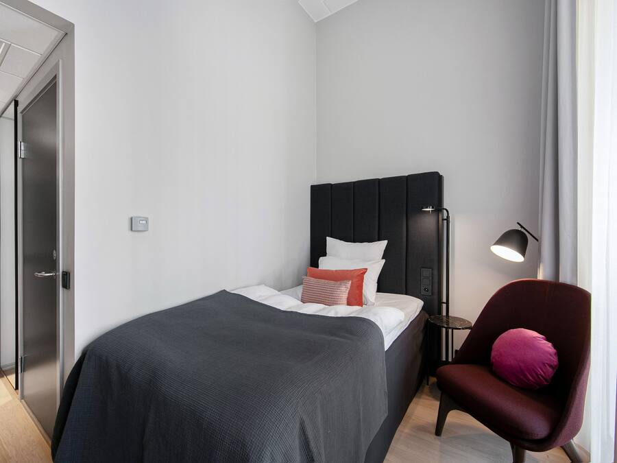 A modern hotel room with a bed, a chair, a bedside table, and a lamp.