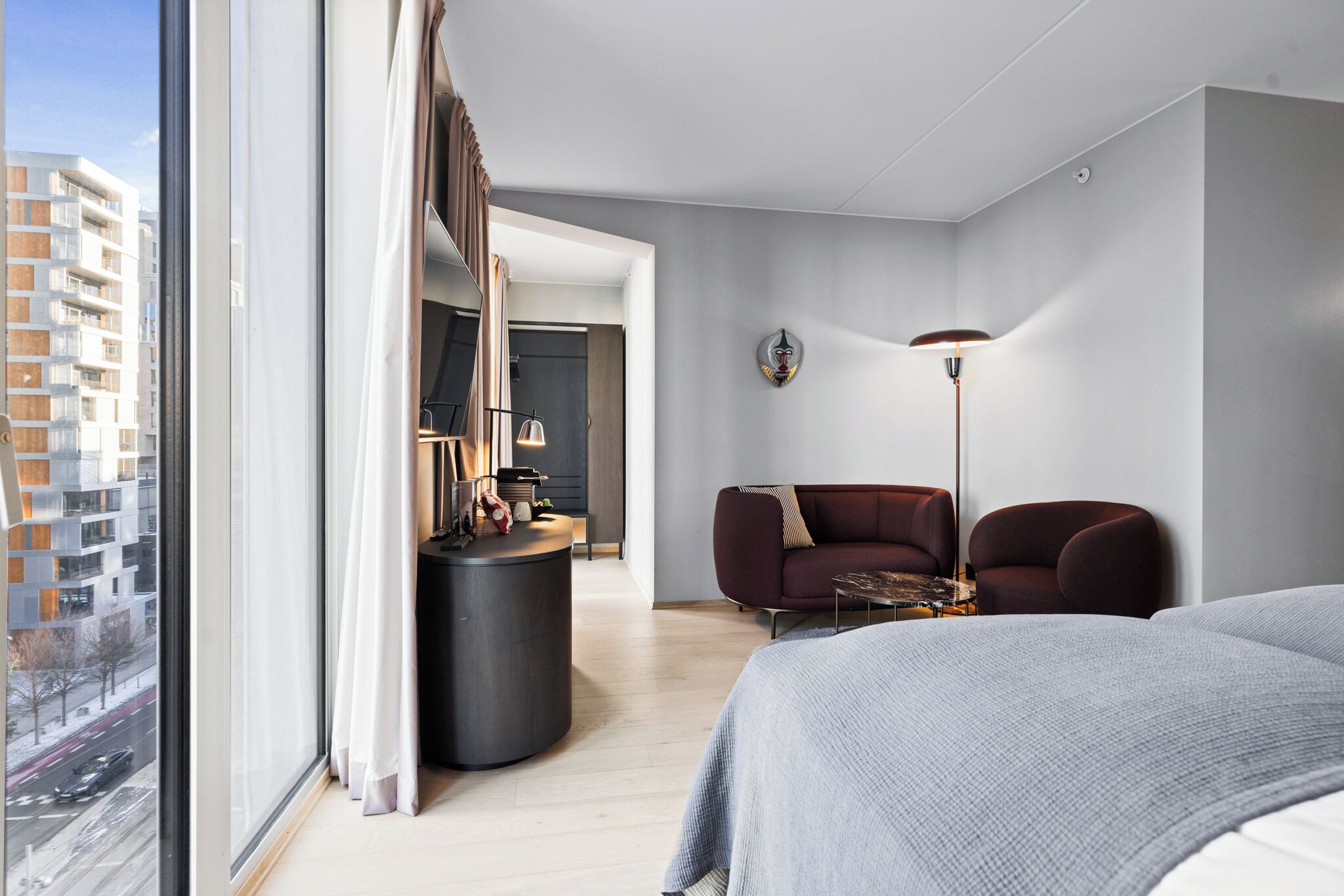 A modern hotel room with a large bed, a seating area with a sofa and armchairs, a small table, a round side table, a floor lamp, and a view of the city through large windows.