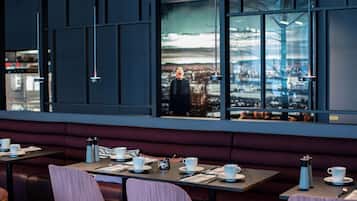 Un restaurant moderne offrant une vue sur la ville, avec des tables décorées de tasses et de soucoupes blanches, et une grande fenêtre qui reflète le paysage urbain.