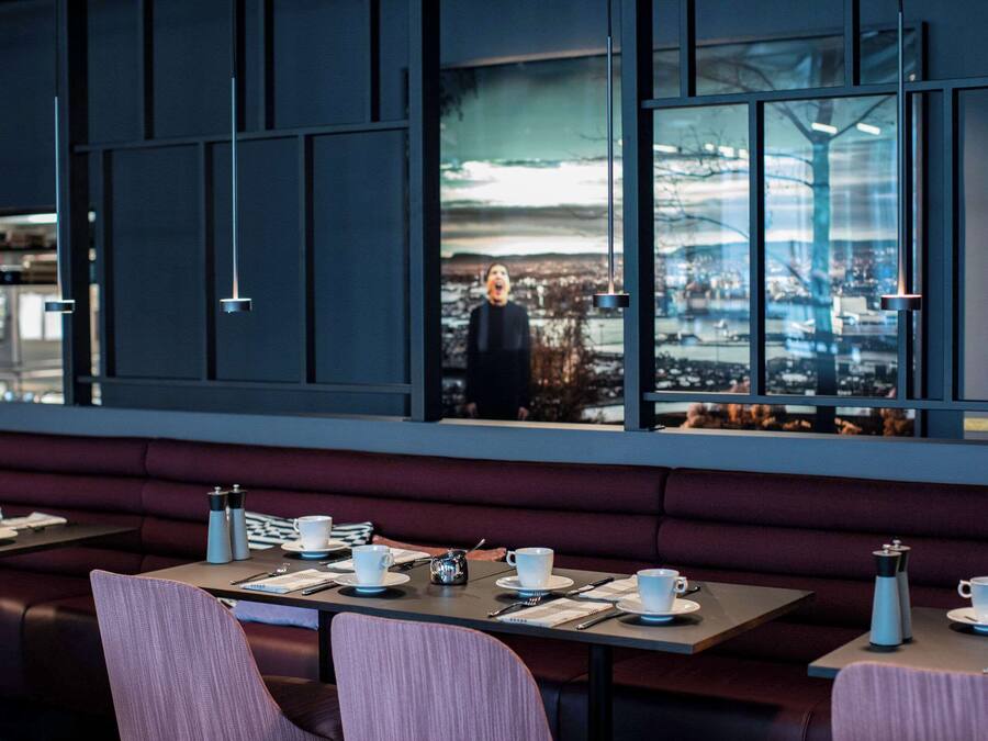 Un restaurant moderne offrant une vue sur la ville, avec des tables décorées de tasses et de soucoupes blanches, et une grande fenêtre qui reflète le paysage urbain.