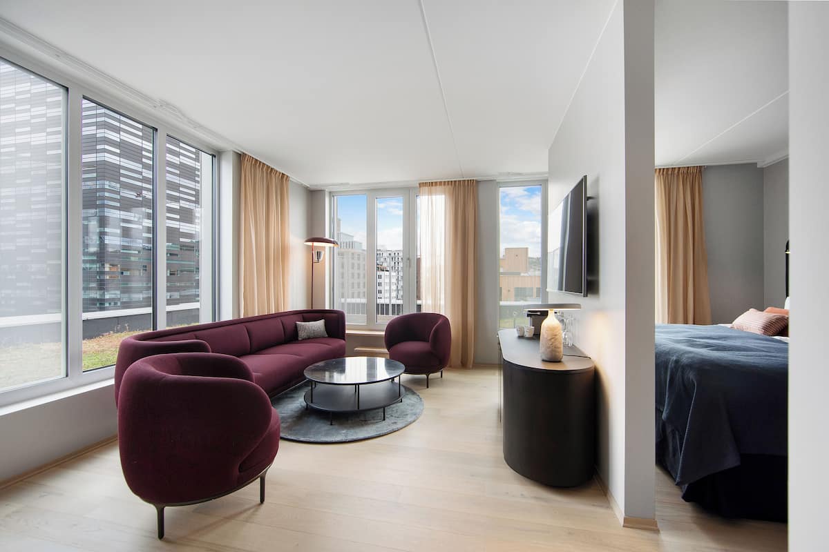 A modern hotel room with large windows, a sofa, a round coffee table, a bed, and a TV.