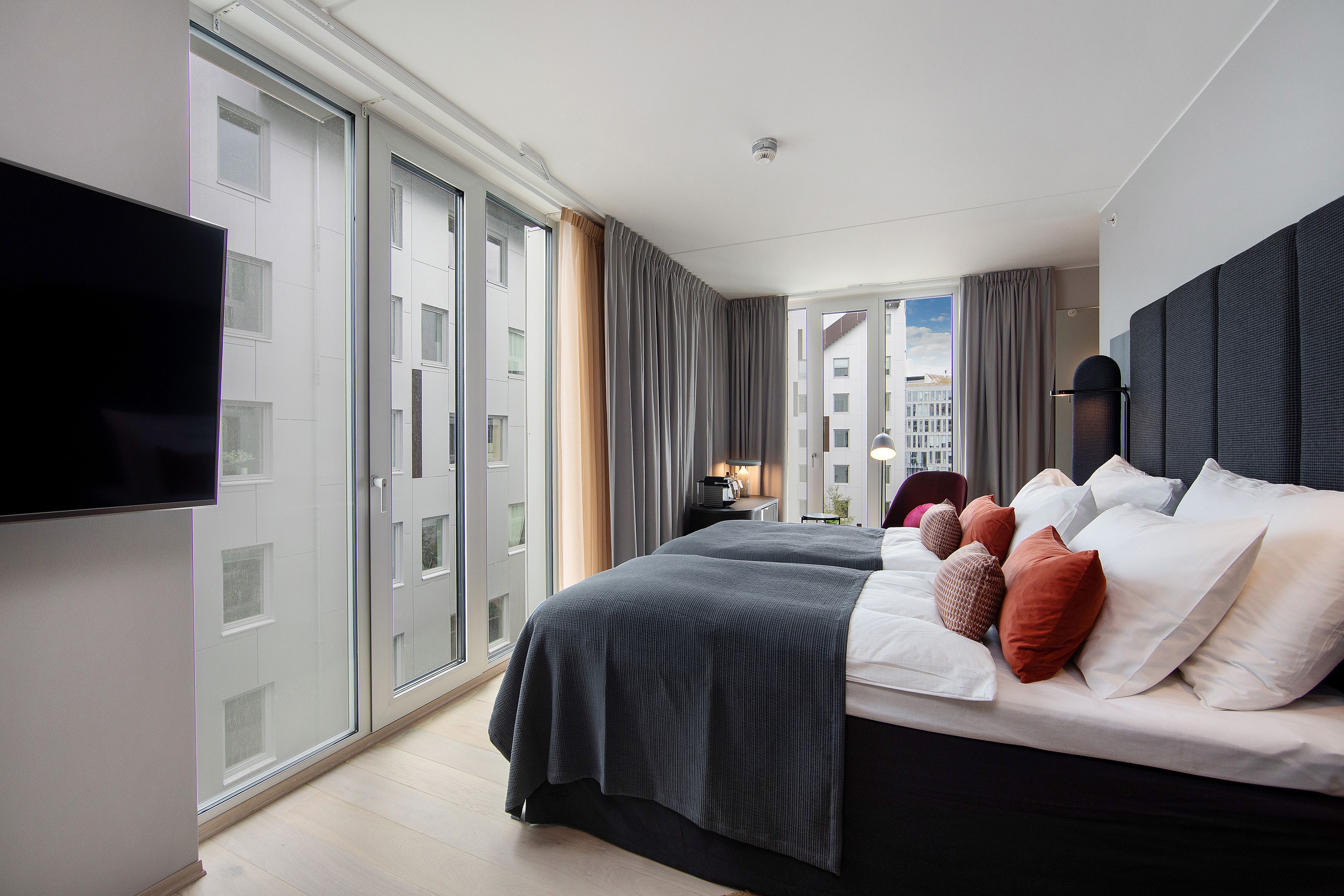 A modern hotel room with a large bed, a flat-screen TV, and a city view through the window.