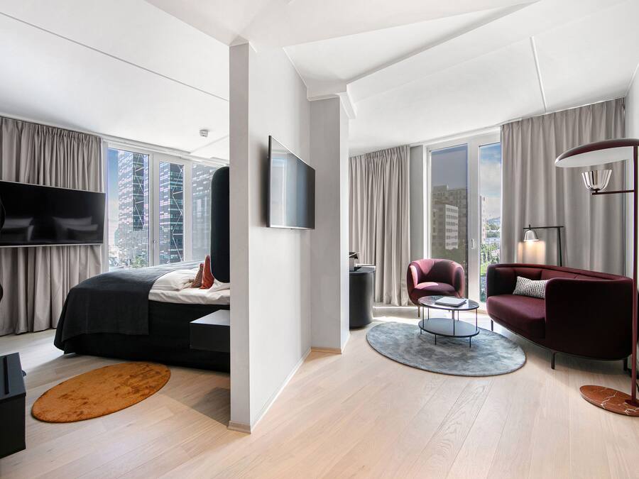 Quarto de hotel moderno com uma cama grande, televisão, área de estar com sofá e poltrona, e amplas janelas com vista para a cidade.