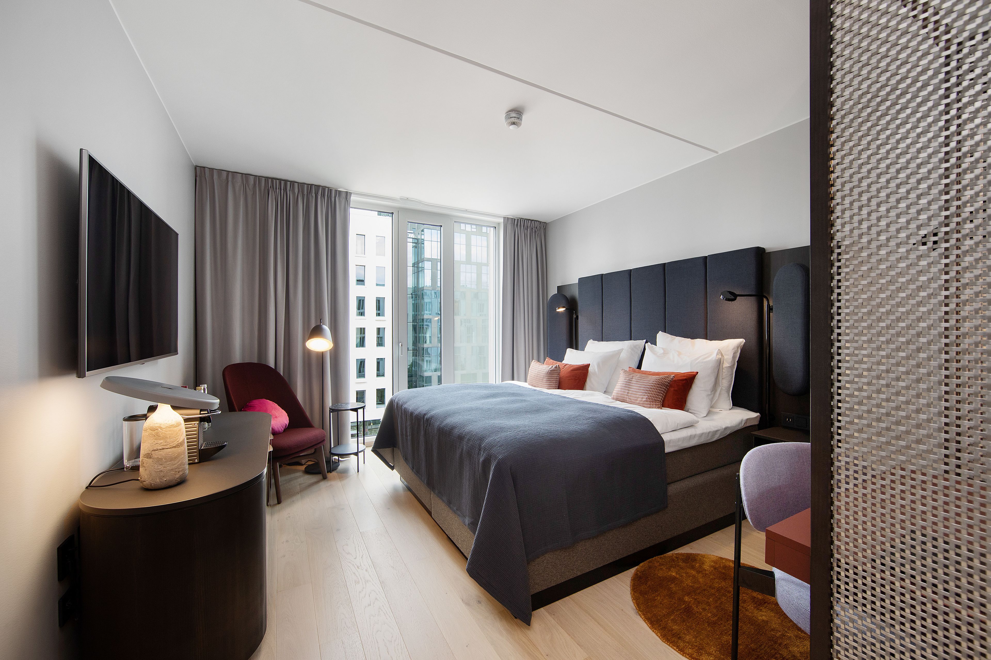 Una habitación de hotel moderna con una cama grande, un escritorio con lámpara, una silla y vistas a la ciudad a través de amplios ventanales.