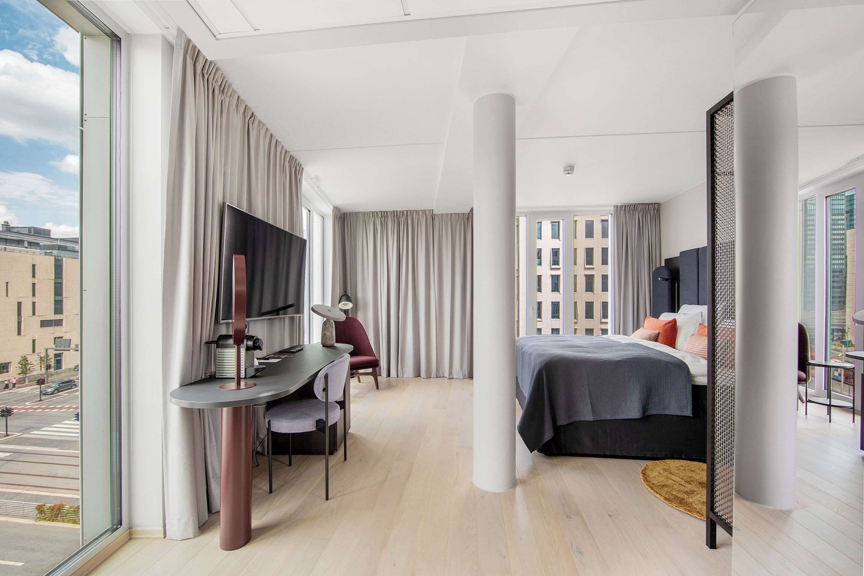 A modern hotel room with a large window, a desk with a chair, a bed with a dark headboard, and a view of the cityscape.