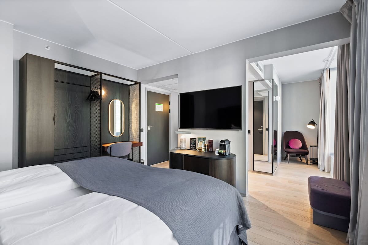 A modern hotel room with a large bed, a desk with a flat-screen TV, a chair, and a small seating area.