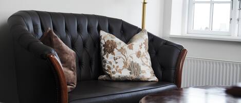 Een donker leren bank met bloemenkussens, een houten bijzettafeltje, een vloerlamp en een raam met jaloezieën.