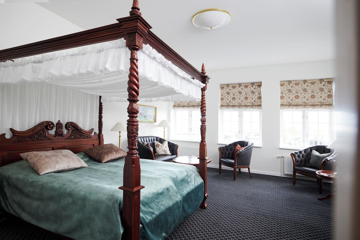 A four-poster bed with a green bedspread, a sitting area with a sofa and armchairs, and a large window with patterned curtains.