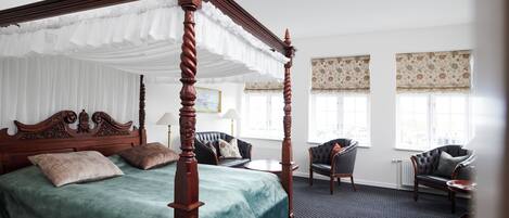 A four-poster bed with a green bedspread, a sitting area with a sofa and armchairs, and a large window with patterned curtains.
