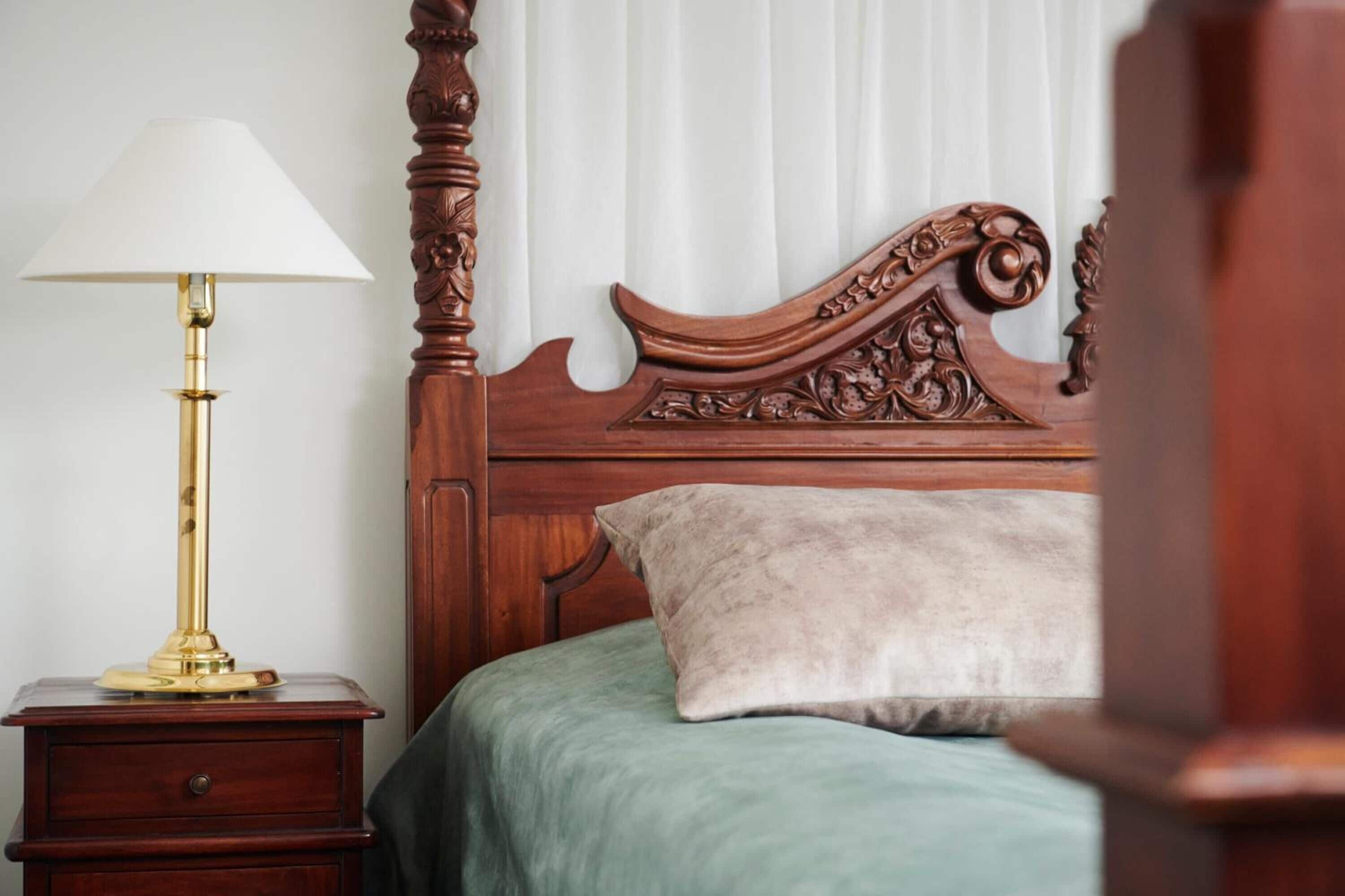 A wooden bed with a green bedspread and a white pillow, a brass table lamp, and a wooden nightstand with a drawer.
