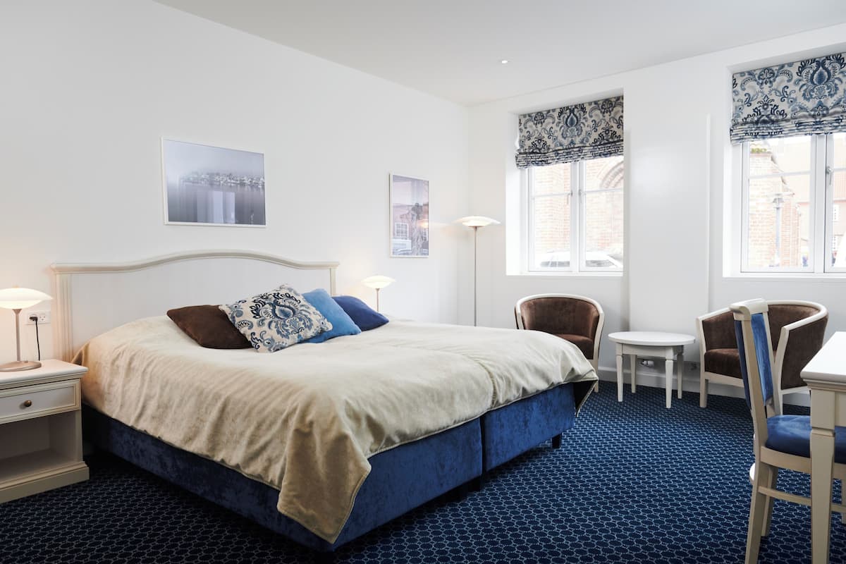 A hotel room with a large bed, a desk, two chairs, a small table, and two windows with patterned curtains.