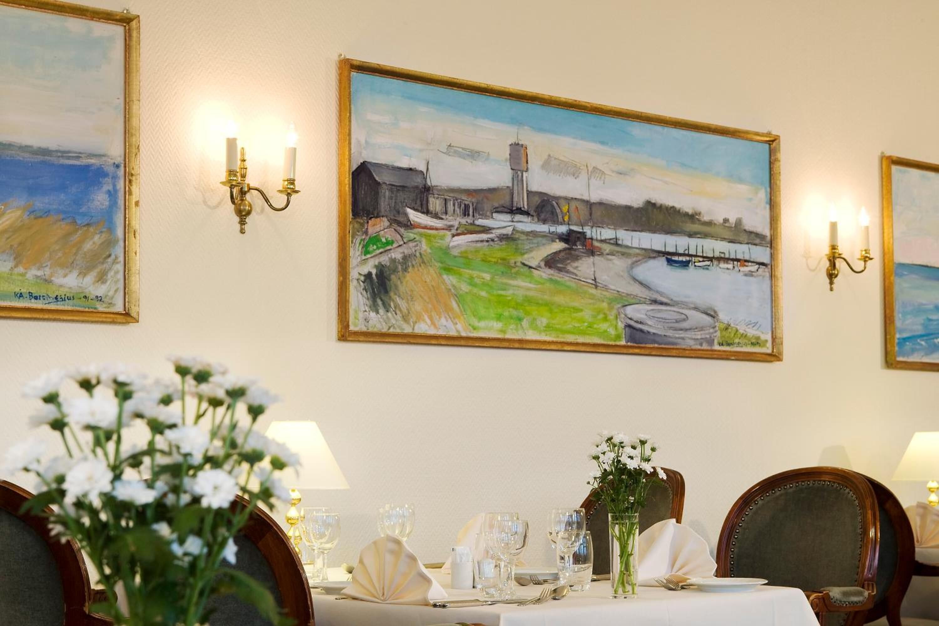 A dining area with a table set for two, framed paintings on the wall, and a vase of flowers on the table.