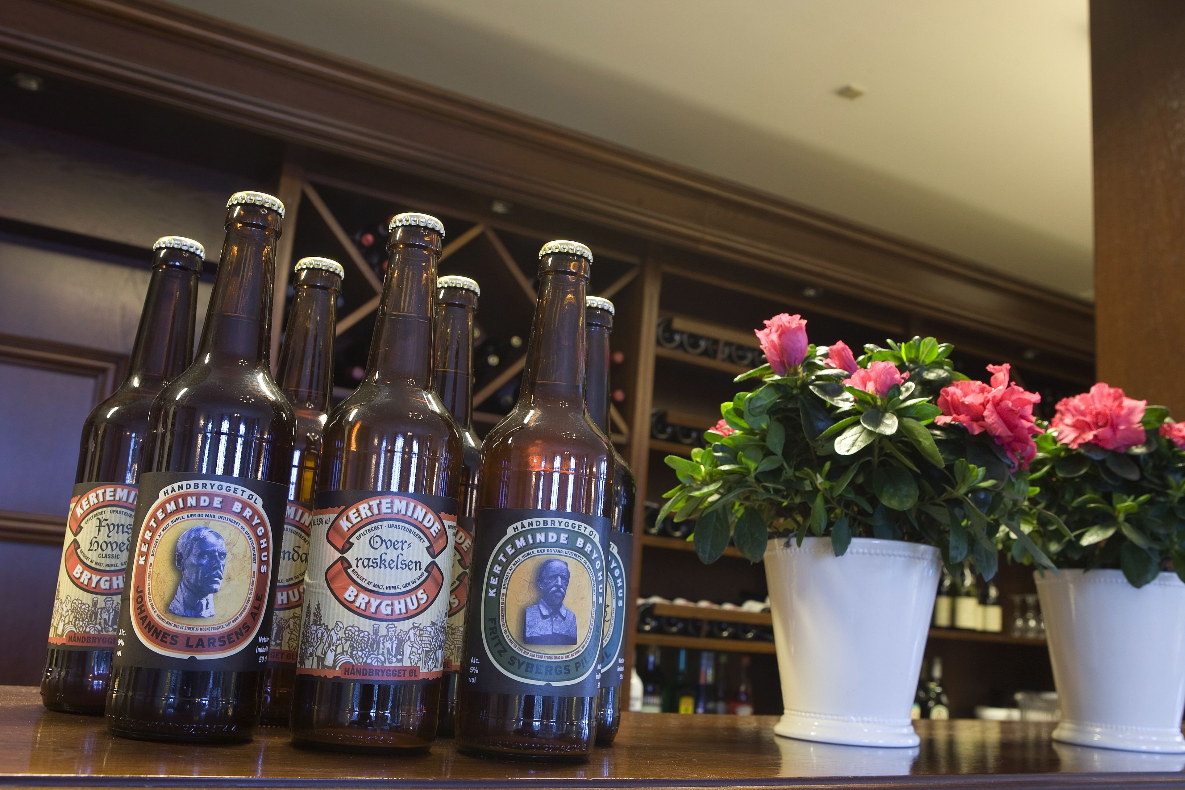 Plusieurs bouteilles de bière avec leurs étiquettes, une plante en pot aux fleurs roses et une étagère en bois.