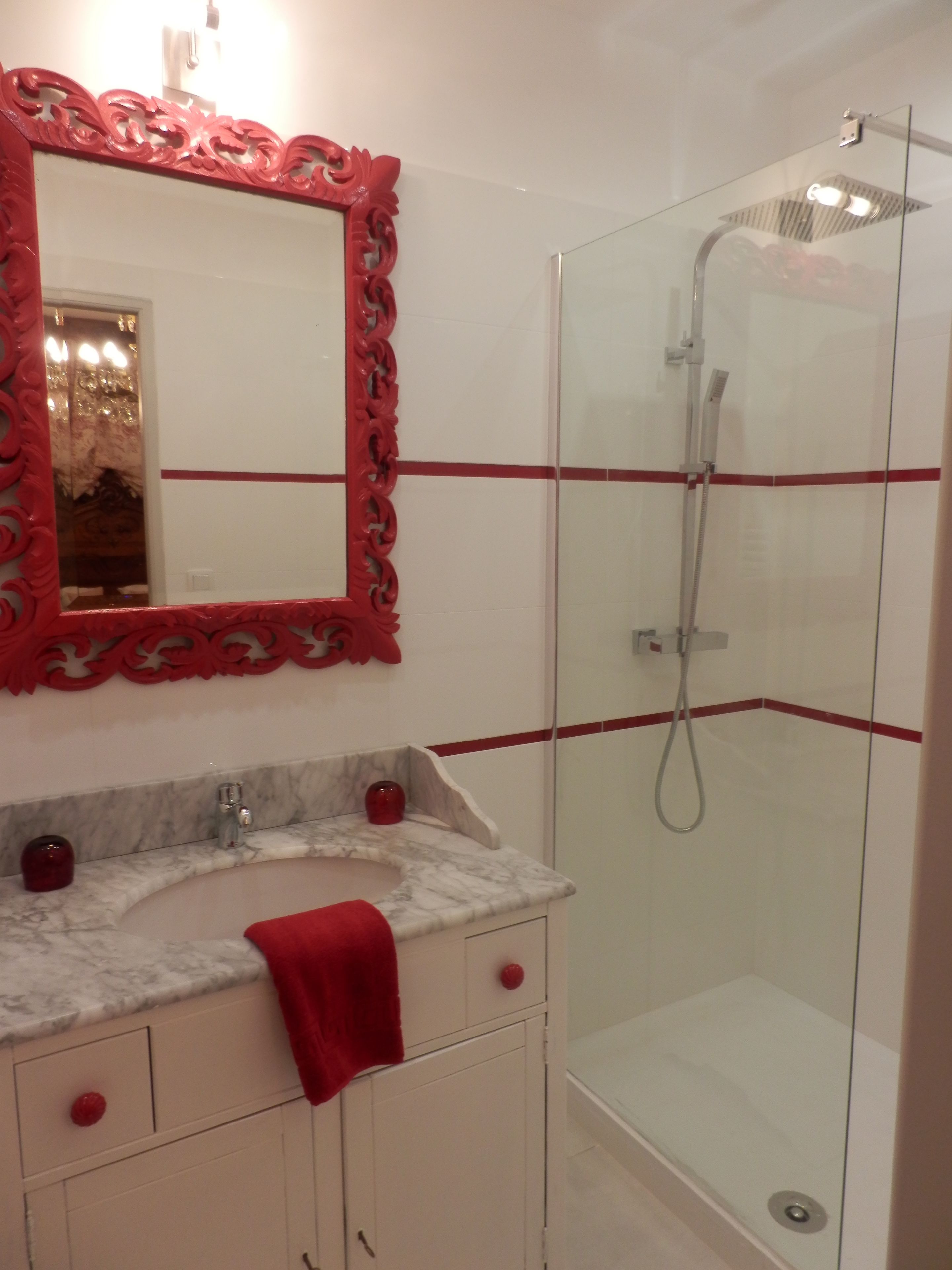 Un baño con lavamanos de mármol, una toalla roja, cabina de ducha de vidrio y un espejo con marco rojo.